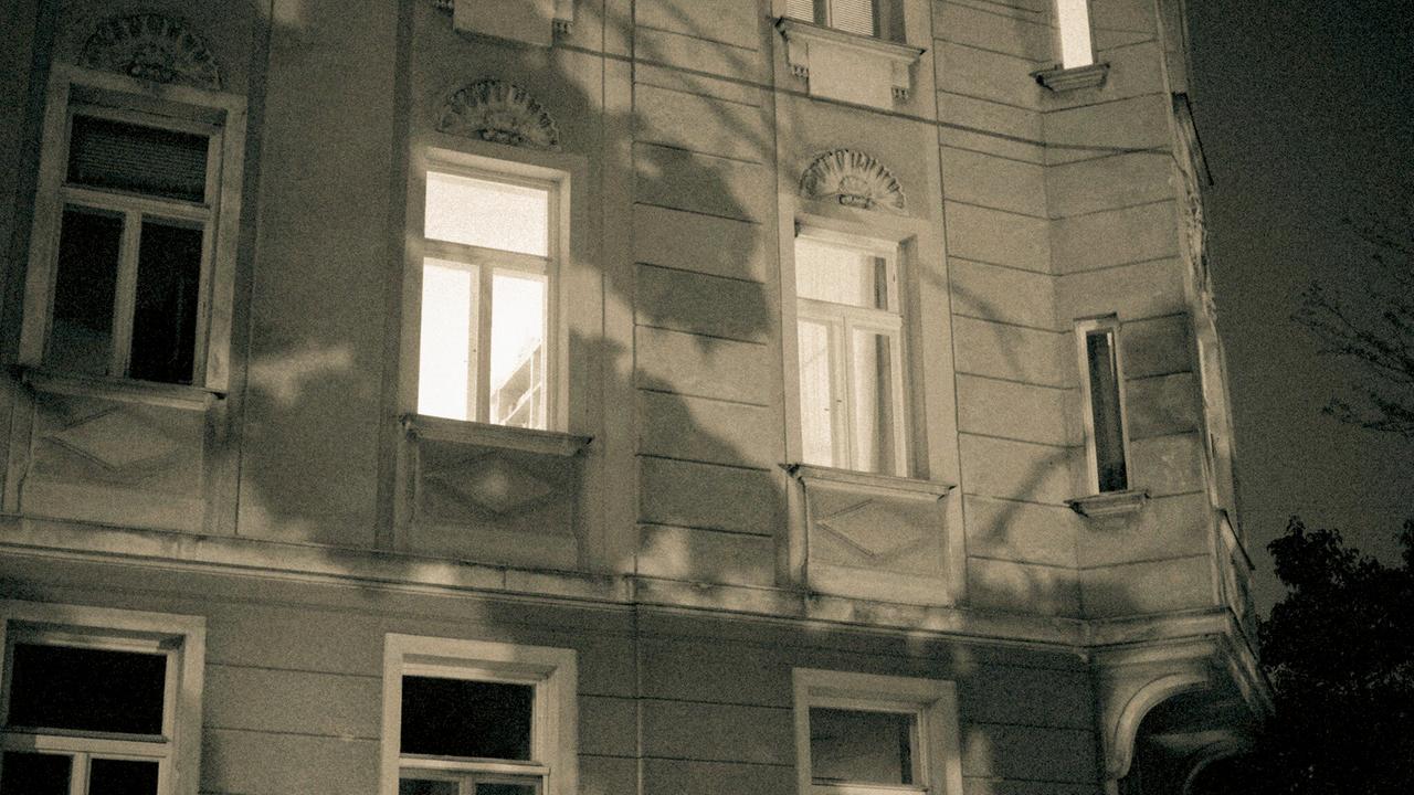 Eine Altbaufassade im Dunkeln, zwei Fenster sind beleuchtet