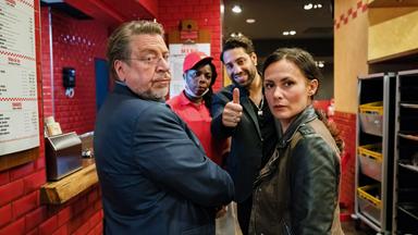 In einem Burger-Restaurant stehen Erichsen (Armin Rohde), Tülay Yildirim (Idil Üner), die Restaurantmanagerin (Dela Dabulamanzi) und Mübariz Pettekaya (Kais Setti) nebeneinander und blicken aufmerksam in eine Richtung.