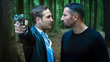 Angelo (Frederick Lau) und Claude (Murathan Muslu) stehen sich angespannt im Wald gegenüber, während Angelo eine Pistole in der ausgestreckten rechten Hand hält.