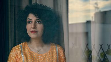 Porträtfoto einer Frau mit dunklem schulterlangem gelocktem Haar, die eine gelbe Bluse trägt und an einem Fenster steht.