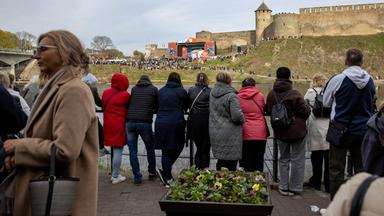 Menschen stehen mit dem Rücken zum Betrachter an einem Geländer und blicken über einen Fluss zum anderen Ufer, wo unterhalb einer Burg eine große Bühne aufgebaut ist, um die sich viele Menschen versammelt haben.