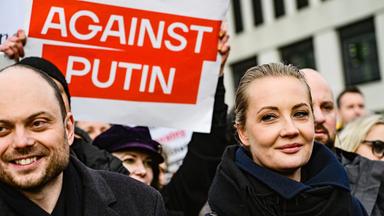 Eine Frau mit streng zurückgekämmten Haaren und bekleidet mit dicker Winterjacke und Schal läuft lächelnd bei einer Demo mit. Hinter ihr hält eine Frau ein rotes Schild mit der Aufschrift Against Putin hoch.
