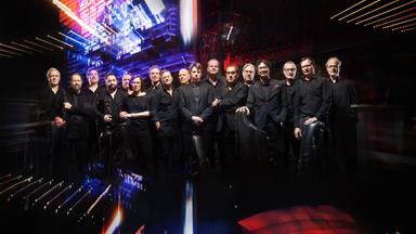 Große Musikgruppe auf einer Bühne mit bunten Lichteffekten; mehrere Musiker mit Instrumenten in Konzertaufstellung.
