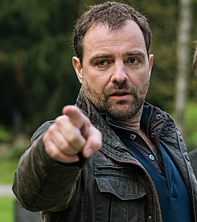 Zwei Männer mittleren Alters in Jacken stehen draußen. Der eine, in einer Lederjacke, zeigt entschlossen mit dem Finger, der andere, in einem Mantel, blickt ernst.