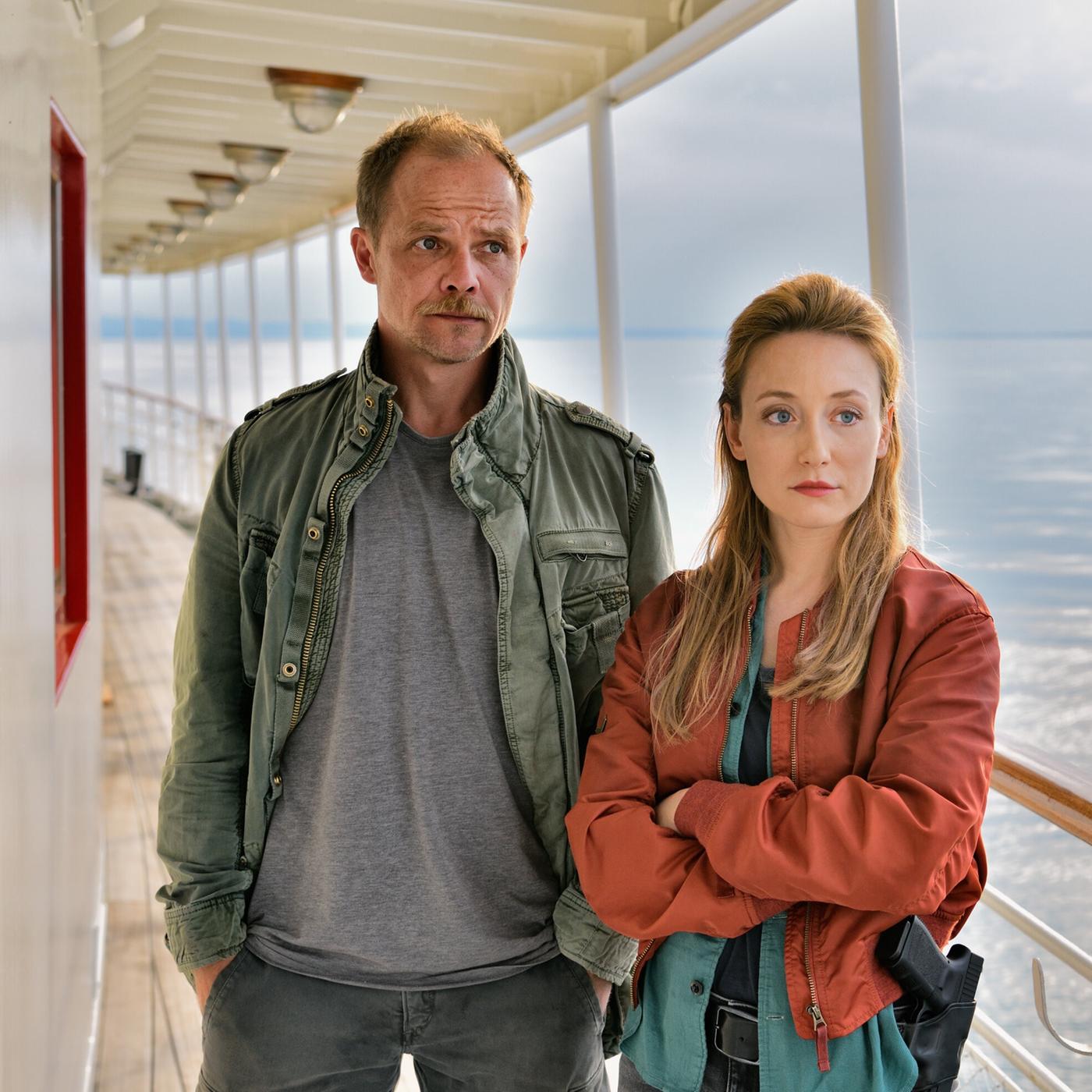 Das Bild zeigt zwei Personen auf einem Schiff. Die Umgebung deutet auf eine maritime Atmosphäre hin, mit einem weiten Wasserbereich im Hintergrund, der von Licht reflektiert wird.   Die erste Person, ein Mann, steht links im Bild. Er trägt eine graue T-Shirt und eine grüne Jacke. Sein Gesicht ist ernst und hat ein leicht nachdenkliches Ausdruck. Die zweite Person, eine Frau, steht rechts. Sie trägt eine rote Jacke über einem blauen Oberteil und hat einen entschlossenen Blick. Beide Personen stehen an einer geschützten Stelle des Schiffs, wo eine Brüstung aus Holz verläuft.  Der Himmel über ihnen wirkt bewölkt, was eine düstere Stimmung vermittelt. Das Bild ist gut beleuchtet, mit einem Fokus auf den Ausdruck der beiden Figuren. Es scheint, als würden sie sich miteinander unterhalten oder eine Beobachtung machen.