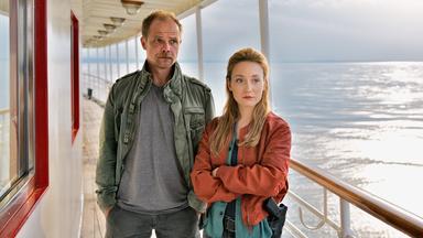 Das Bild zeigt zwei Personen auf einem Schiff. Die Umgebung deutet auf eine maritime Atmosphäre hin, mit einem weiten Wasserbereich im Hintergrund, der von Licht reflektiert wird.   Die erste Person, ein Mann, steht links im Bild. Er trägt eine graue T-Shirt und eine grüne Jacke. Sein Gesicht ist ernst und hat ein leicht nachdenkliches Ausdruck. Die zweite Person, eine Frau, steht rechts. Sie trägt eine rote Jacke über einem blauen Oberteil und hat einen entschlossenen Blick. Beide Personen stehen an einer geschützten Stelle des Schiffs, wo eine Brüstung aus Holz verläuft.  Der Himmel über ihnen wirkt bewölkt, was eine düstere Stimmung vermittelt. Das Bild ist gut beleuchtet, mit einem Fokus auf den Ausdruck der beiden Figuren. Es scheint, als würden sie sich miteinander unterhalten oder eine Beobachtung machen.