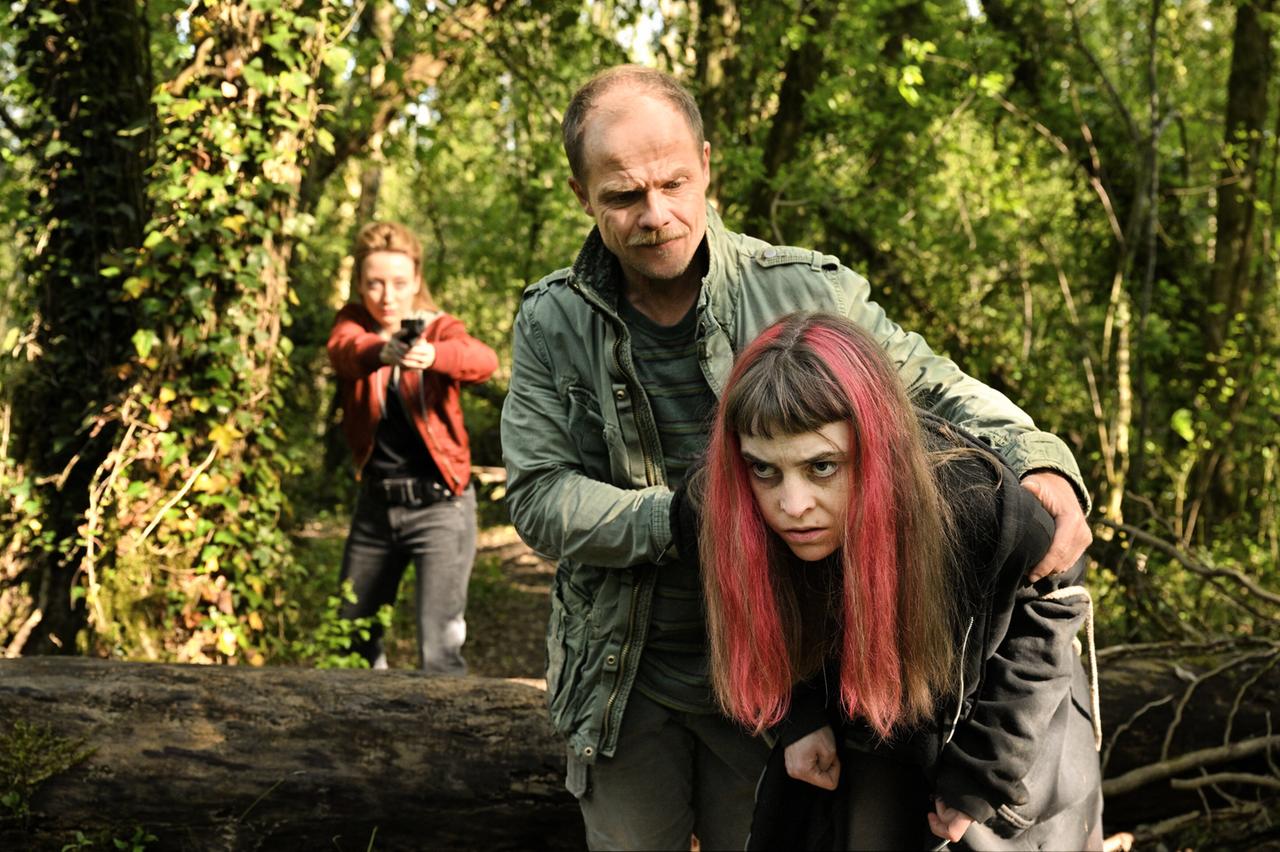 Das Bild zeigt eine Szene aus einem Wald. Im Vordergrund stehen drei Personen: Ein Mann hält eine Frau, die in dunkler Kleidung mit langen, braunen Haaren und pinken Strähnen gekleidet ist. Der Mann hat ein ernstes Gesicht und trägt eine olivgrüne Jacke.   Hinter ihnen steht eine zweite Frau in einem roten Oberteil, die eine schwarze Pistole in Richtung der anderen zwei Personen im Vordergrund hält. Die Umgebung ist geprägt von dichter, grüner Vegetation sowie Bäumen, die im Hintergrund sichtbar sind. Der Boden besteht aus einem Baumstamm, der quer im Bild verläuft. Die gesamte Szenerie vermittelt eine angespannte Atmosphäre.