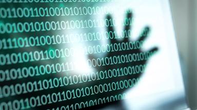 Ein Monitor mit einer Zahlenreihe, bestehend aus 0 und 1 und die Spiegelung einer Hand, die danach greift