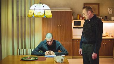 Ein junger Mann sitzt an einem Tisch und schaut konzentriert auf ein Blatt Papier, während ein älterer Mann steht und auf ihn herabblickt. Die Szene spielt in einer schlicht eingerichteten Küche, beleuchtet von einer hängenden Lampe.
