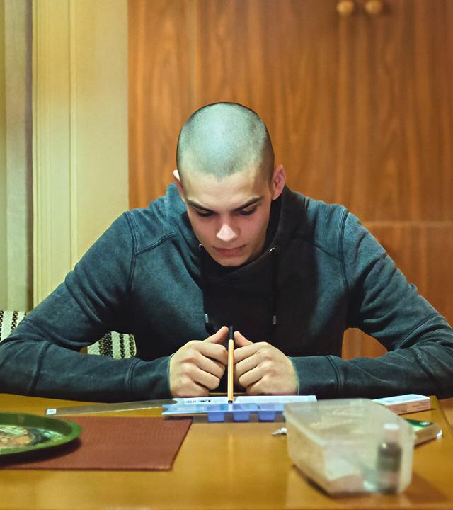 Ein junger Mann sitzt an einem Tisch und schaut konzentriert auf ein Blatt Papier, während ein älterer Mann steht und auf ihn herabblickt. Die Szene spielt in einer schlicht eingerichteten Küche, beleuchtet von einer hängenden Lampe.