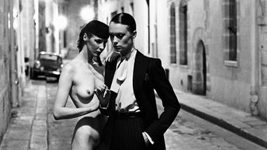 Auf dem schwarz-weiß Bild sieht man zwei Frauen nebeneinander in einer kleinen Straße stehen, es ist Nacht. Die eine trägt einen Anzug, High Heels, Hemd und Jacke, die andere ist nackt bis auf High Heels und einen Hut mit Netzschleier.