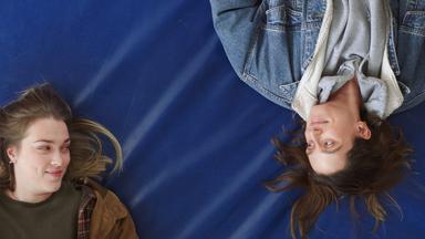 Man schaut von oben auf zwei junge Frauen, die auf einer blauen Turnmatte liegen und sich anschauen. Eine von ihnen hält ein Buch in den Händen.