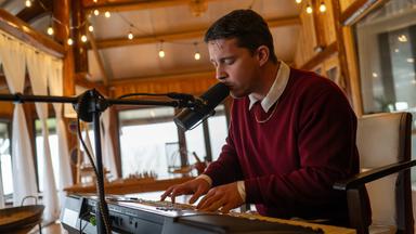 Nico Santos sitzt und spielt auf einem Keyboard und singt dabei in ein Mikrofon, das vor ihm steht. 