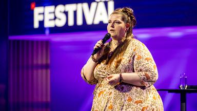 Kabarettistin teht in der Mitte des Bildes. Sie blickt zum linken Bildrand. In ihrer rechten Hand hält sie ein Mikrofon. Sie trägt ein gemustertes Kleid. Die Bühne ist blau und lila beleuchtet.