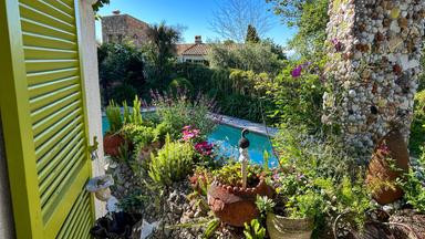 Das Bild zeigt einen Garten mit einer hellen, freundlichen Atmosphäre. Im Vordergrund ist eine grüne Fensterläden-Tür geöffnet, die einen Blick auf eine bunte Gartenlandschaft ermöglicht.   Rechts vom Bild ist eine Säule, die mit Muscheln und Steinen verziert ist. Um diese Säule herum wachsen verschiedene Pflanzen, darunter bunte Blumen in Töpfen aus Ton und Keramik sowie Blattpflanzen. In der Nähe steht ein rustikaler, metallener Stuhl.  Im Hintergrund ist ein Swimmingpool sichtbar, der von üppiger grüner Vegetation umgeben ist. Die Sonne scheint klar, und der Himmel ist strahlend blau, was zur lebhaften Stimmung des Gartens beiträgt.   Insgesamt vermittelt das Bild eine friedliche und einladende Gartenlandschaft an der Côte d'Azur in Südfrankreich.