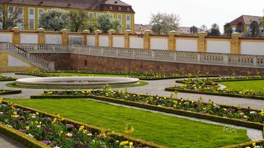 Das Bild zeigt eine wunderschöne Gartenlandschaft, die mit bunten Blumen und grünen Rasenflächen gestaltet ist. Im Vordergrund sind rechteckige Blumenbeete mit verschiedenen Blumenarten, darunter Tulpen und andere Farbtupfer, angeordnet. Diese Beete sind von frisch geschnittenem Rasen umgeben.  Im Hintergrund ist eine große, gelbe Gebäudeanlage zu sehen, die von höherem, geschnittenem Grün flankiert wird. Ein flacher, runder Brunnen aus hellem Stein ist ebenfalls im Bild vorhanden, umgeben von einer geschwungenen Pflasterung. Auf der linken Seite führen Treppen zu einer höher gelegenen Mauer, die mit balustradenartigen Ausstattungen verziert ist. Die gesamte Szenerie vermittelt eine harmonische und gepflegte Gartenatmosphäre. Es ist ein klarer, sonniger Tag, und die Blütenfärbungen heben sich lebhaft vom Grün des Gartens ab.
