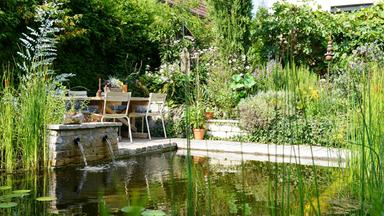 Naturgarten mit Sitzplatz auf einer kleinen Stein-Terrasse am Teich, üppige Bepflanzung aus Blumen, Kräutern und Sträuchern, Wasserfontänen im Vordergrund