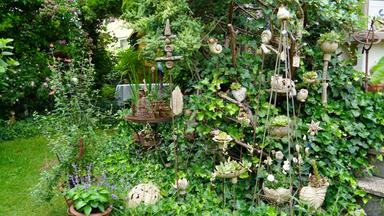 Der Garten zeigt eine vielfältige Bepflanzung mit alten Gehölzen, Rosen, Beerensträuchern und Blühpflanzen. Eine Garten-Modellbahn verläuft durch die Pflanzen. Dekorative Töpfe und Figuren sind ebenfalls sichtbar.