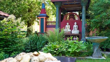 Das Bild zeigt einen naturnahen Garten mit asiatischen Einflüssen. Im Vordergrund sind Pflanzen und Holzobjekte, im Hintergrund stehen zwei Buddha-Statuen vor einer farbigen Wand. Ein Brunnen ist ebenfalls sichtbar.