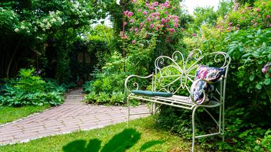 Das Bild zeigt einen Garten mit üppiger Vegetation. Im Vordergrund steht eine helle, schmiedeeiserne Bank mit einem dicken, dunkelgrünen Polster und einem dekorativen Kissen mit floralem Muster. Um die Bank herum sind verschiedene Pflanzen angeordnet, darunter große grüne Blätter und Sträucher, die farbige Blüten tragen, insbesondere rosafarbene Blüten. Der Weg, der durch den Garten führt, besteht aus dunkelroten, rechteckigen Steinen und windet sich durch die dichten Pflanzen. Im Hintergrund sind hohe Bäume und Sträucher zu erkennen, die einen natürlichen und ruhigen Ambiente schaffen. Der Garten ist sehr bepflanzt und vermittelt ein Gefühl von Verwilderung und gleichzeitigem Design.
