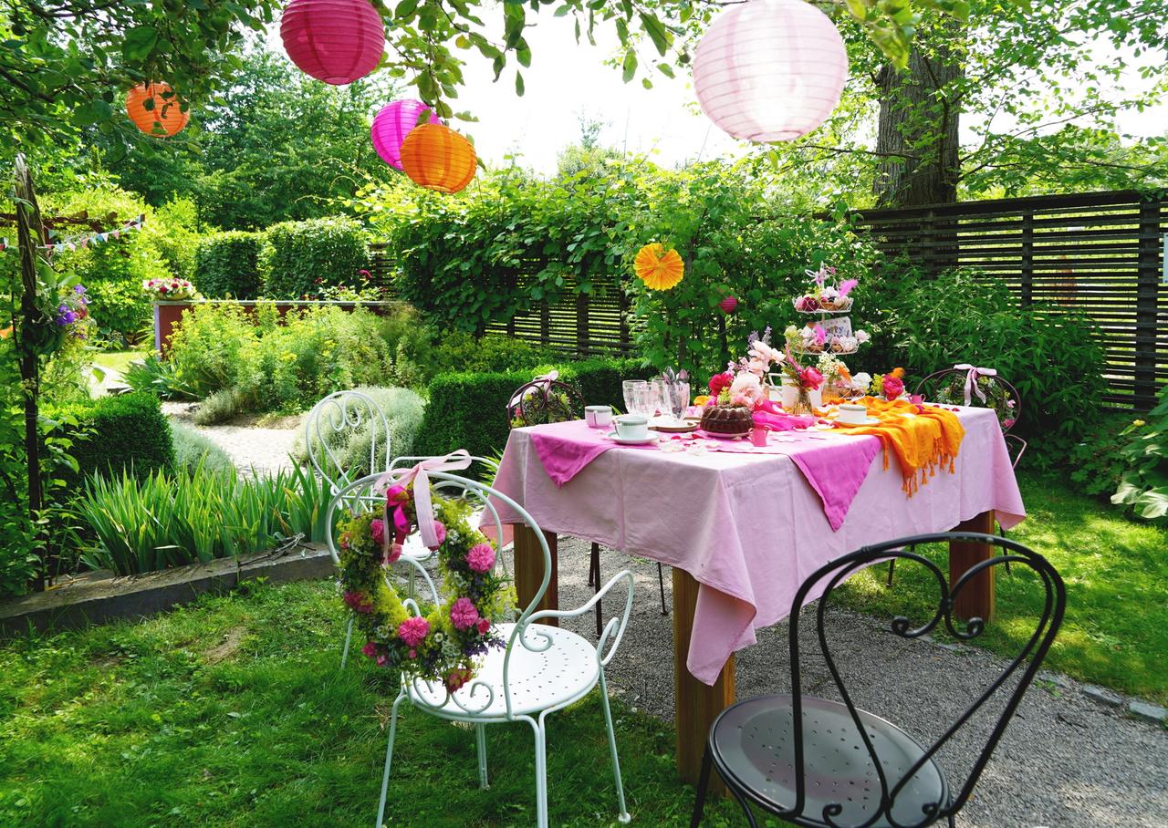 Festlich gedeckter Tisch mit Blumen, Gebäck und bunten Laternen in einem Garten.  