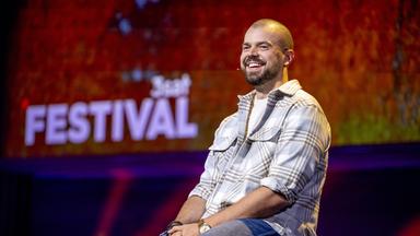 Nikita Miller sitzt auf einem Barhocker in der Mitte des Bildes. Er trägt ein offenes kariertes Hemd und lächelt. Der Hintergrund ist rot beleuchtet.