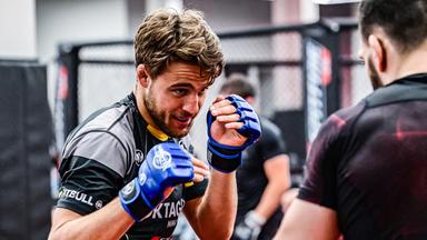 Ein junger Mann mit Bart, in Trainingskleidung mit Schutzhandschuhen, kämpft in einem Studio gegen einen Gegner. 