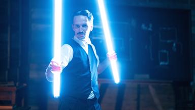 Spielszene: Nikola Tesla steht in seiner Werkstatt mit leuchtenden Röhren, die er wie Schwerter in der Hand hält.