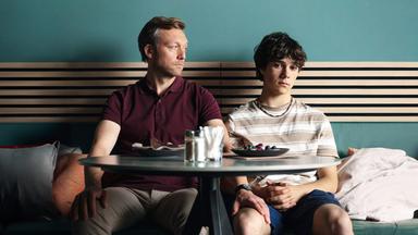 Gabo (Carlo Krammling) und Dave (Robin Sondermann) sitzen nebeneinander an einem Cafétisch. Dave sieht zu Gabo und hat seine Hand auf dessen Oberschenkel gelegt. Gabo schaut gedankenverloren vor sich hin.