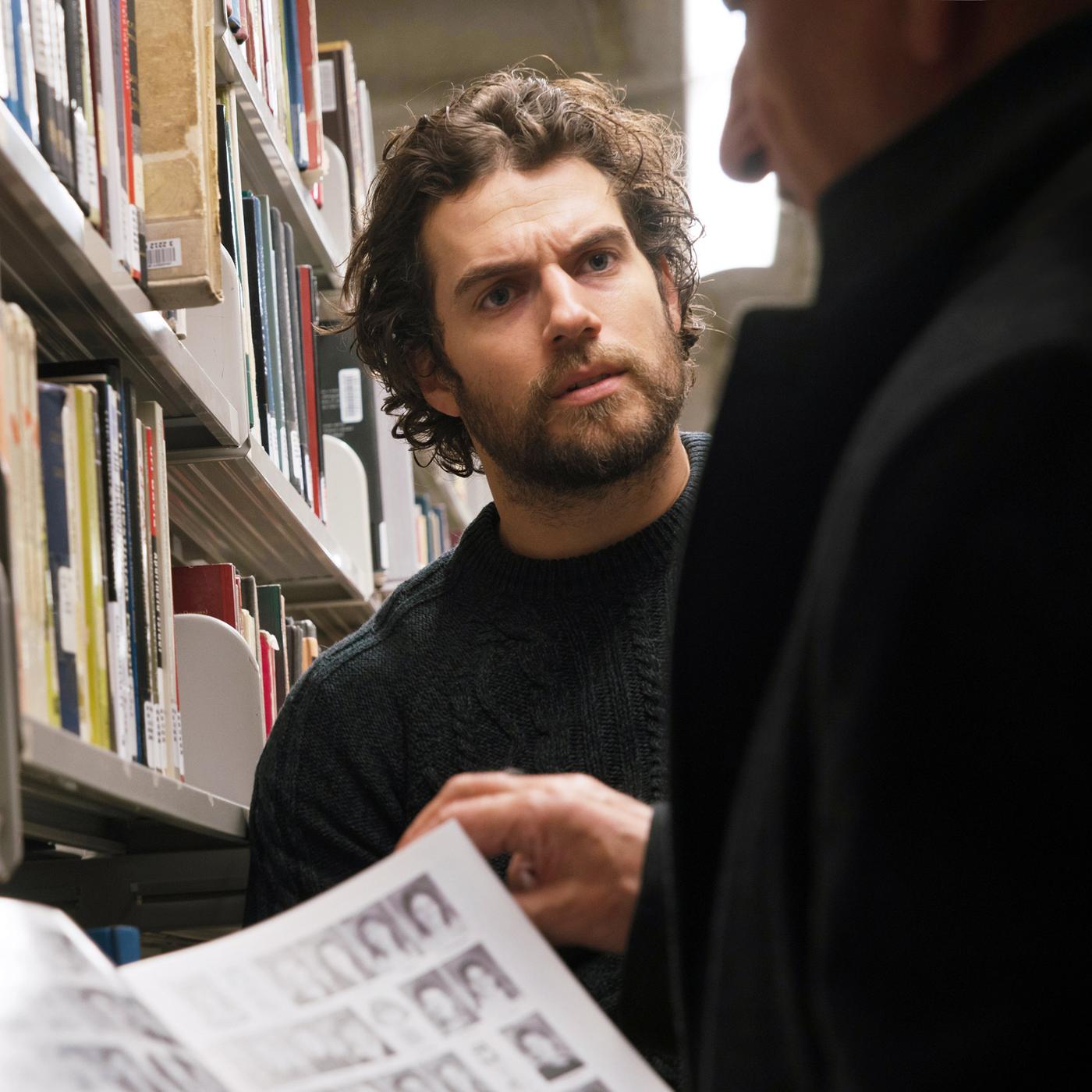 ooper (Ben Kingsley) und Marshall (Henry Cavill) stehen in einer Bibliothek. Cooper ist nur leicht im Profil zu sehen und hält eine Zeitschrift geöffnet vor sich. Marshall sieht ihn mit ernstem Blick an.