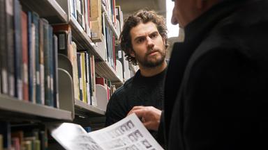 ooper (Ben Kingsley) und Marshall (Henry Cavill) stehen in einer Bibliothek. Cooper ist nur leicht im Profil zu sehen und hält eine Zeitschrift geöffnet vor sich. Marshall sieht ihn mit ernstem Blick an.