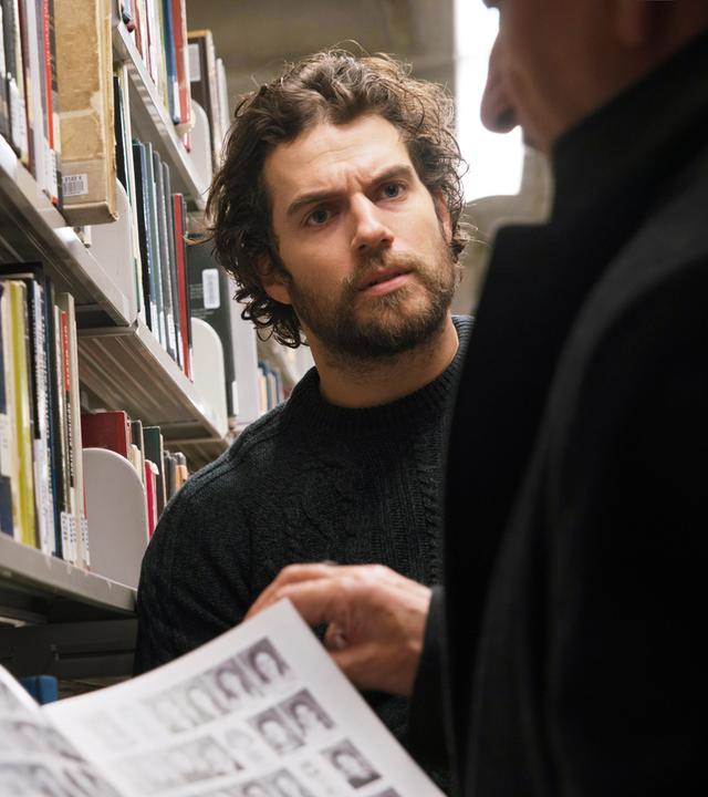 ooper (Ben Kingsley) und Marshall (Henry Cavill) stehen in einer Bibliothek. Cooper ist nur leicht im Profil zu sehen und hält eine Zeitschrift geöffnet vor sich. Marshall sieht ihn mit ernstem Blick an.