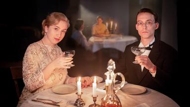 Szene aus "Reichskanzlerplatz" von Nora Bossong. An einem eingedeckten Tisch mit weißer Tischdecke sitzen eine Frau und ein Mann. Sie halten ein gefülltes Weinglas in der Hand. 