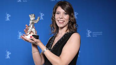 EIne Frau mit braunen Haaren und schwarzem Kleid steht vor einer Wand mit Berlinale-Logos und hält lächelnd einen Preis in Form eines silberenen Bären in die Kamera.