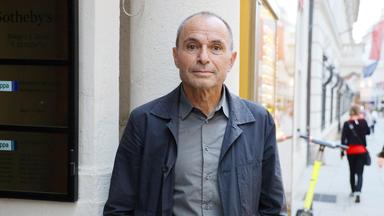 Ein älterer Mann imd unkelgrauen Anzug steht an einer Straßenecke.