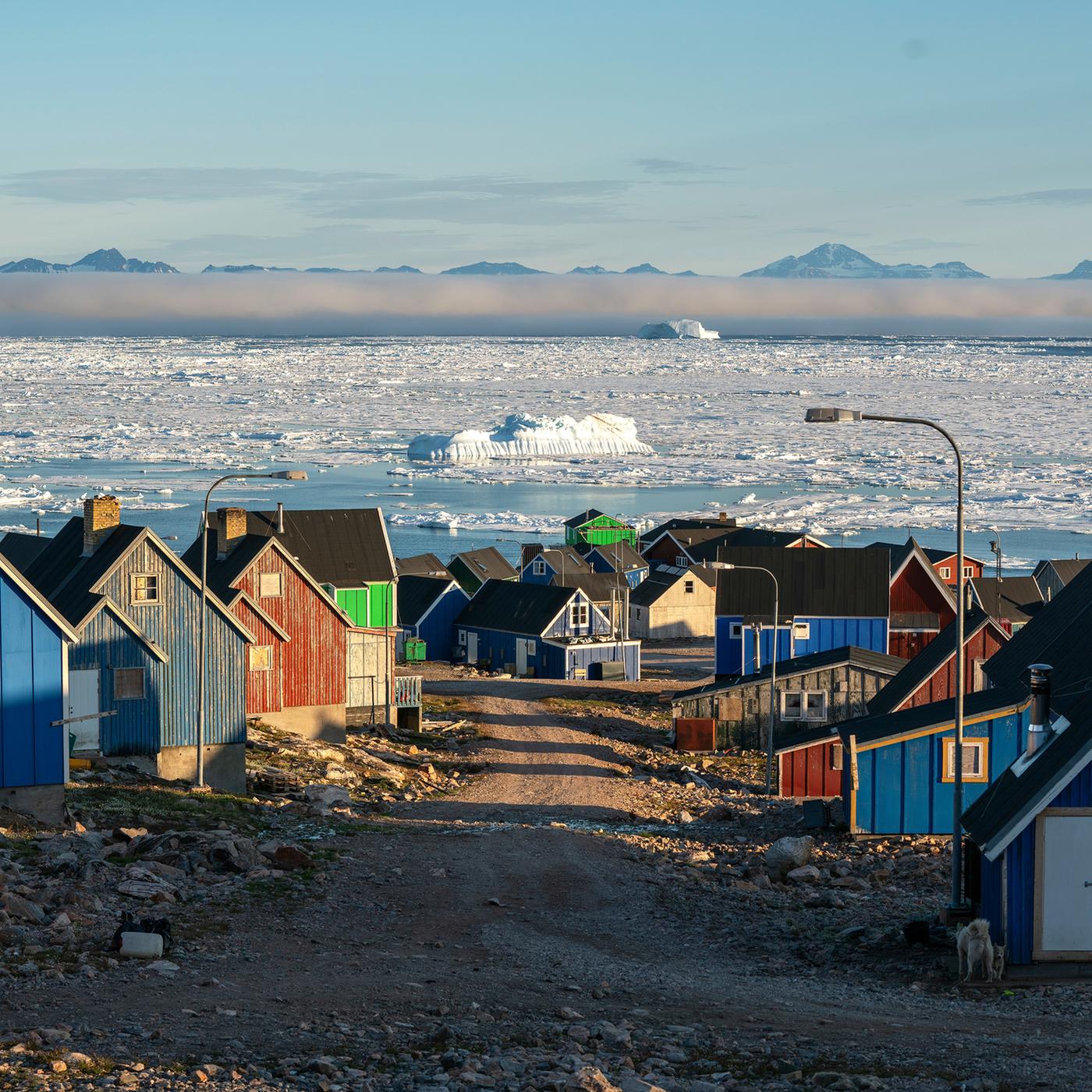 Bunte Holzhäuser getrennt druch eine Schotterstraße, im Hintergrund Eismeer mit von Nebel bedeckten Bergen.