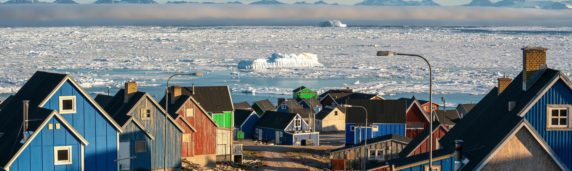 Bunte Holzhäuser getrennt druch eine Schotterstraße, im Hintergrund Eismeer mit von Nebel bedeckten Bergen.