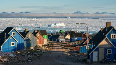 Bunte Holzhäuser getrennt druch eine Schotterstraße, im Hintergrund Eismeer mit von Nebel bedeckten Bergen.