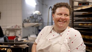 Eine Frau steht mit einer Kochschürze in einer Backstube. Das T-Shirt, welches sie darunter trägt, ist mit kleinen Herzen bedruckt.