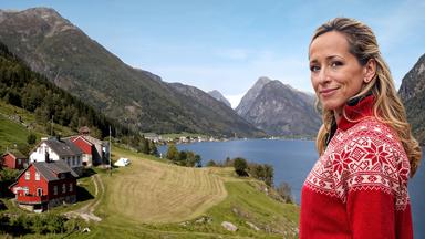 Eine blonde Frau vor einer Landschaft mit Fjord, Bergen und Wiesen.