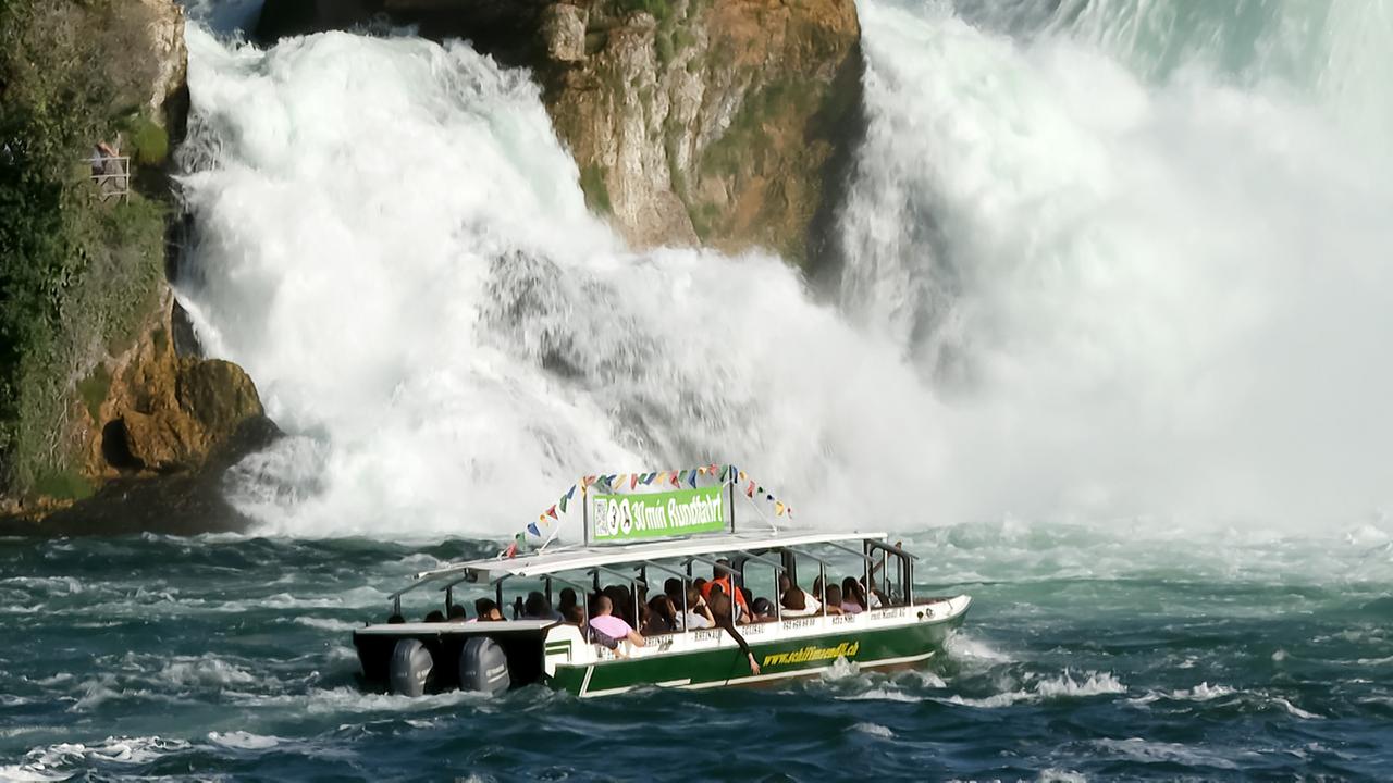 Das Bild zeigt ein Boot mit Passagieren, das im Wasser nahe den Rheinfällen fährt. Im Hintergrund sind rauschende Wasserfälle sowie grüne Felsen sichtbar.