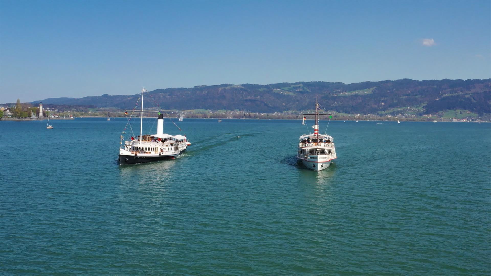 Im Bild sind zwei historische Schiffe auf dem Bodensee zu sehen. Der Schaufelraddampfer Hohentwiel und das Schiff MS Österreich fahren nebeneinander in ruhigem Wasser. Im Hintergrund sind sanfte Hügel sichtbar.
