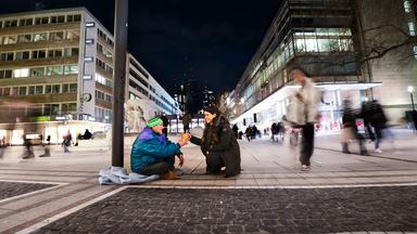 Es ist Nacht in der Frankfurter Fußgängerzone. Im Vordergrund sitzt ein obdachloser Mann mit grüner Mütze. Vor ihm kniet eine junge Frau mit langen schwarzen Haaren, die dem Obdachlosen einen Mahlzeit überreicht.
