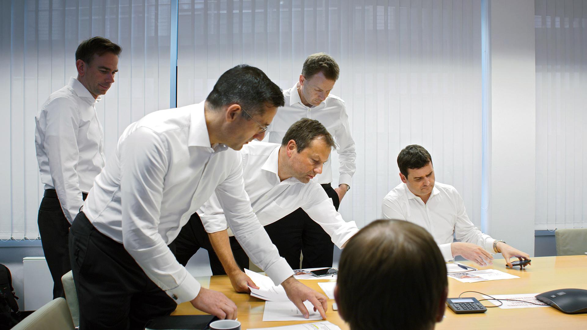 Um einen Tisch herum stehen vier Männer und sitzen zwei weitere und beugen sich über ausgebreitete Papiere. Alle Männer tragen ein weißes Hemd und eine schwarze Hose. Der oberste Hemdknopf ist geöffnet. Alle sind bartlos und haben kurze Haare. Sie wirken konzentriert. Vor die Fenster sind Lamellen gezogen.