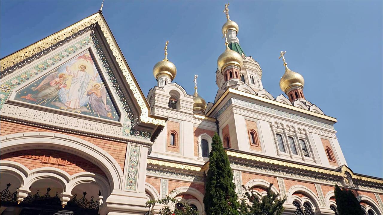 Das Bild zeigt eine russische Kirche mit goldenen Kuppeln. Der Teil der Kirche auf der linken Seite hat ein dekoratives Mosaik und kunstvolle Verzierungen. Der Himmel ist klar und blau.