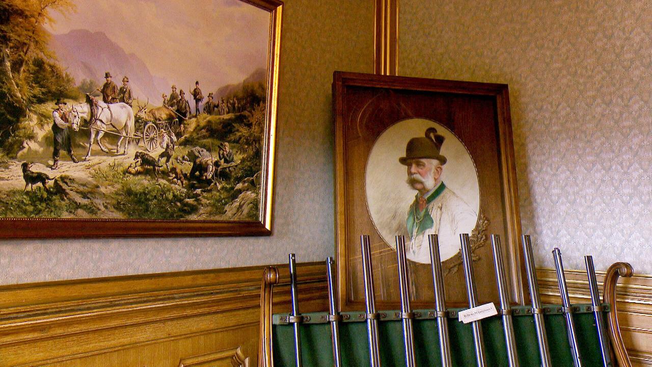 Zwei alte Bilder an der Wand vor denen diverse Gewehre angelehnt stehen.