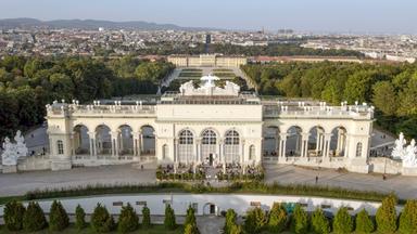 Das Bild zeigt die Gloriette, ein architektonisches Bauwerk im Schönbrunner Schlosspark in Wien, Österreich. Die Gloriette ist auf einem erhöhten Platz errichtet und bietet einen weiten Blick über die Stadt Wien und die umliegenden Landschaften.   Im Vordergrund sind die beeindruckenden Säulen und Bögen der Gloriette zu sehen, die in einem hellen Farbton gehalten sind. Die Fassade ist reich verziert, und über dem Hauptteil des Gebäudes befinden sich Skulpturen, darunter figürliche Darstellungen.  Unterhalb der Gloriette sind Besucher zu erkennen, die auf einer Terrasse sitzen und die Aussicht genießen. Im Hintergrund zieht sich die Stadt Wien mit ihren charakteristischen Gebäuden und Bäumen in die Ferne. Der Himmel ist klar, was eine helle und freundliche Atmosphäre schafft.   Die gesamte Szene vermittelt ein Gefühl von historischer Eleganz und natürlicher Schönheit, umgeben von der Pracht der österreichischen Landschaft.