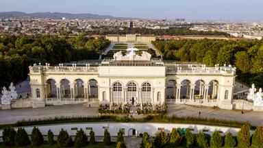 Das Bild zeigt die Gloriette, ein architektonisches Bauwerk im Schönbrunner Schlosspark in Wien, Österreich. Die Gloriette ist auf einem erhöhten Platz errichtet und bietet einen weiten Blick über die Stadt Wien und die umliegenden Landschaften.   Im Vordergrund sind die beeindruckenden Säulen und Bögen der Gloriette zu sehen, die in einem hellen Farbton gehalten sind. Die Fassade ist reich verziert, und über dem Hauptteil des Gebäudes befinden sich Skulpturen, darunter figürliche Darstellungen.  Unterhalb der Gloriette sind Besucher zu erkennen, die auf einer Terrasse sitzen und die Aussicht genießen. Im Hintergrund zieht sich die Stadt Wien mit ihren charakteristischen Gebäuden und Bäumen in die Ferne. Der Himmel ist klar, was eine helle und freundliche Atmosphäre schafft.   Die gesamte Szene vermittelt ein Gefühl von historischer Eleganz und natürlicher Schönheit, umgeben von der Pracht der österreichischen Landschaft.