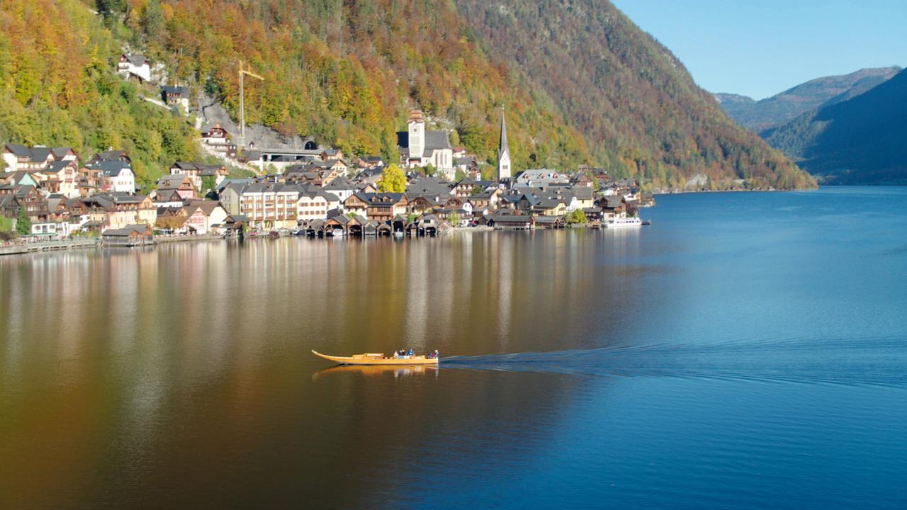 Das Bild zeigt eine malerische Landschaft am Hallstättersee in Österreich, mit dem charmanten Dorf Hallstatt im Hintergrund. Die Szenerie ist geprägt von sanften Hügeln, die mit buntem Laub bewachsen sind, und einem klaren blauen Himmel. In der Mitte des Wassers bewegt sich ein langes, schmalen Boot mit mehreren Personen, das sanfte Wellen hinterlässt. Am Ufer sind diverse Häuser und Gebäude sichtbar, darunter traditionelle österreichische Chalets mit dunklen Holzverkleidungen und bunten Fassaden. In der Nähe des Dorfes sind auch einige Boote, die an einem Steg festgemacht sind. Im Hintergrund ragt eine Kirchturmspitze empor, umgeben von steilen Bergen. Das Wasser reflektiert die Umgebung und trägt zur ruhigen, friedlichen Atmosphäre bei.