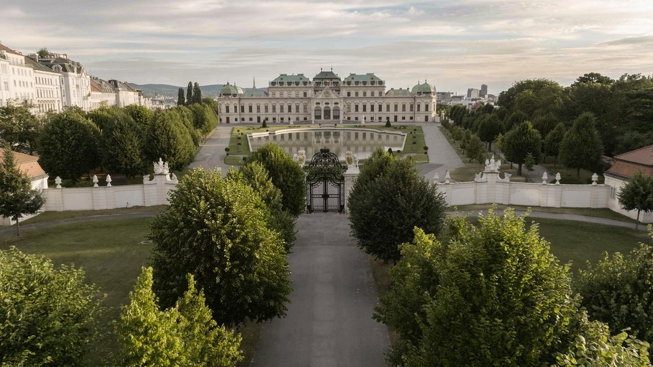 Das Bild zeigt das Obere Belvedere in Wien, ein prachtvolles barockes Schloss. Im Vordergrund verläuft ein gepflasterter Weg, flankiert von hohen, grünen Bäumen. Der Weg führt zu einem schmiedeeisernen Tor, das den Zugang zu einem gepflegten Garten markiert. Hinter dem Garten erstreckt sich ein größerer, rechteckiger Teich, dessen Oberfläche das Licht reflektiert.   Das Schloss selbst ist eindrucksvoll mit einer symmetrischen Fassade gestaltet, die von eleganten Fenstern und einladenden Eingangstüren geprägt ist. Die Dächer sind mit grünen Kupferblechen gedeckt und haben eine markante Form. In der Umgebung sind weitere Bäume sowie Bauten im Hintergrund zu sehen, die das städtische Ambiente der Stadt Wien betonen. Der Himmel ist teils bewölkt, was dem Bild eine ruhige und friedliche Atmosphäre verleiht.